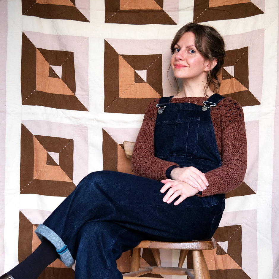 Woman sitting on a stool wearing blue overalls and a brown sweater against a geometric-patterned wall.