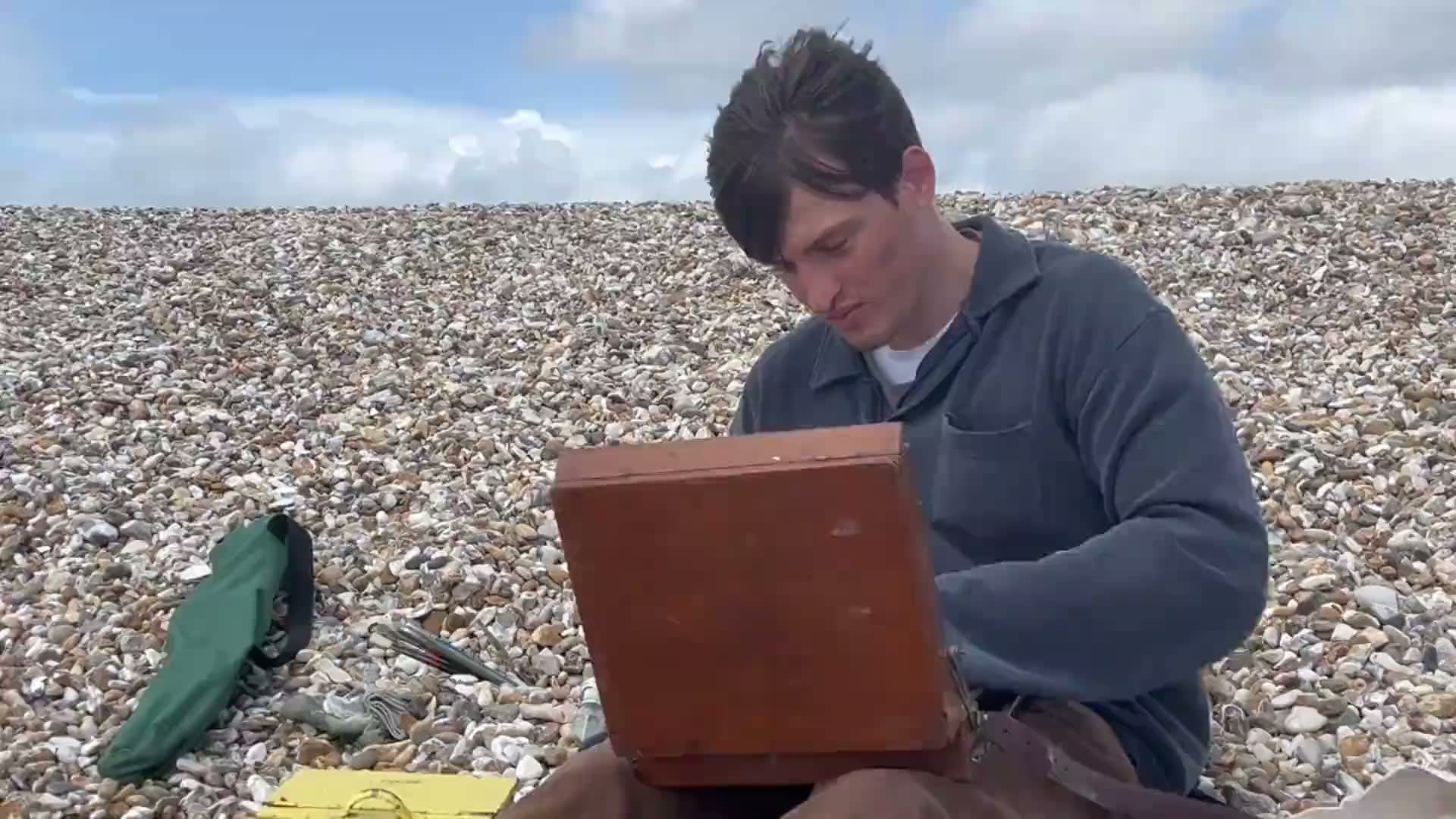 Joshua Lea painting on a shingle beach, with his paint box open beside him